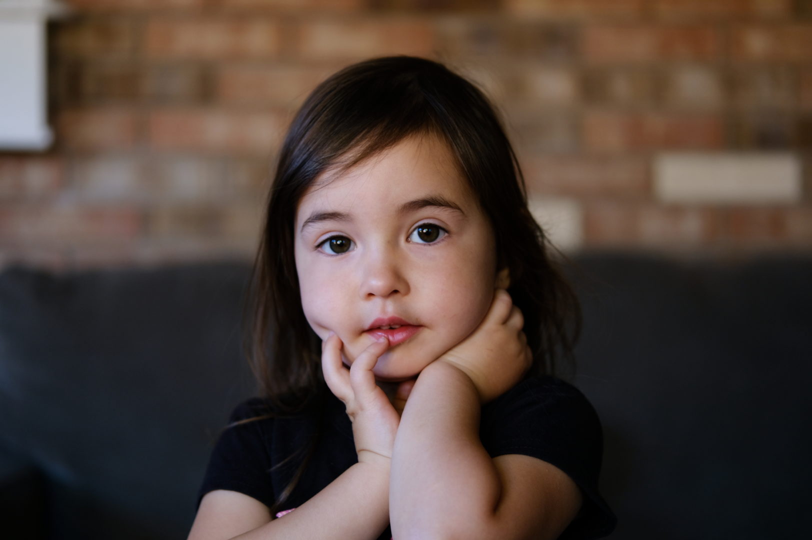A 2 year old girl poses for the camera against a brick wall.
