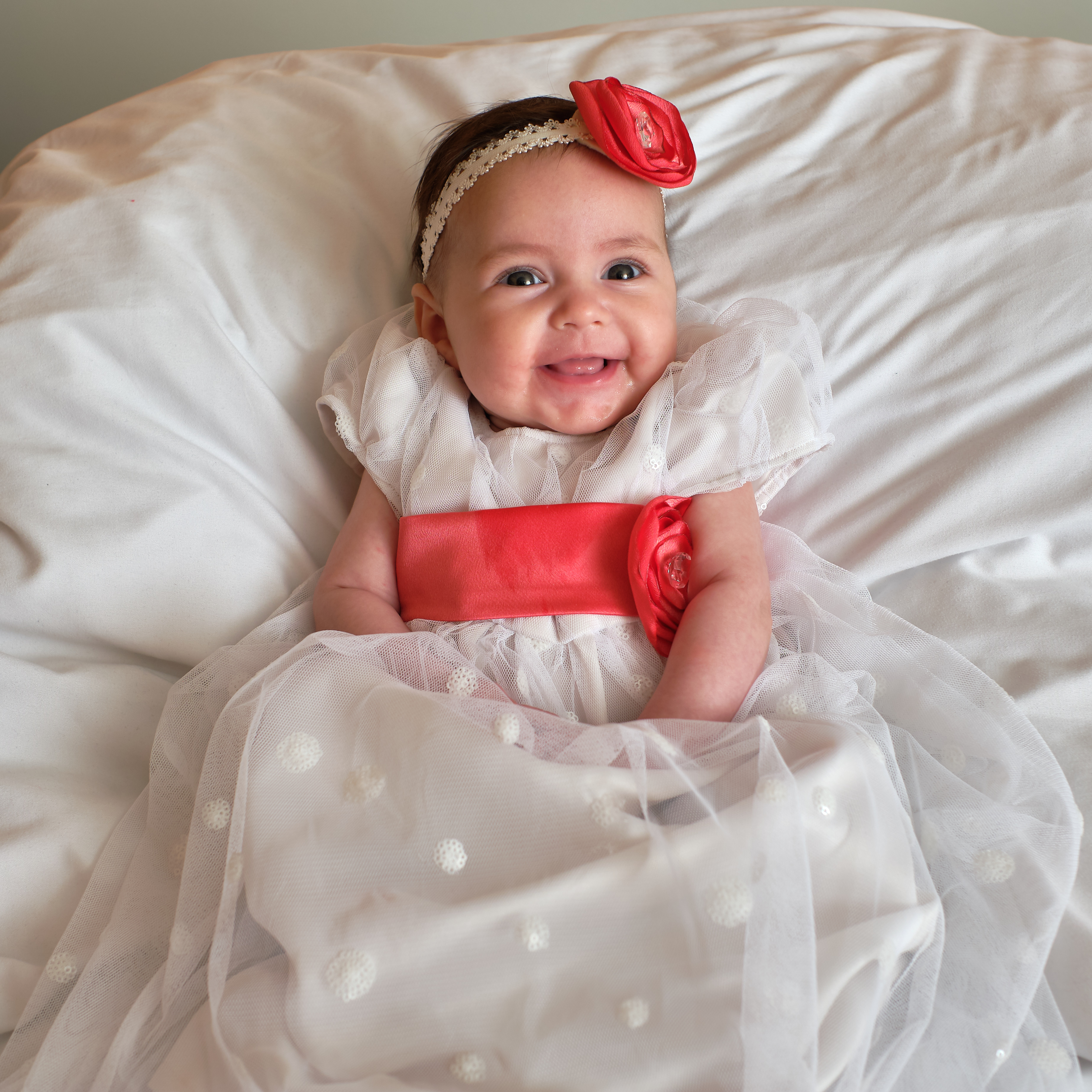 Baby Emily in a long white dress with a red sash and a red bow in her hair