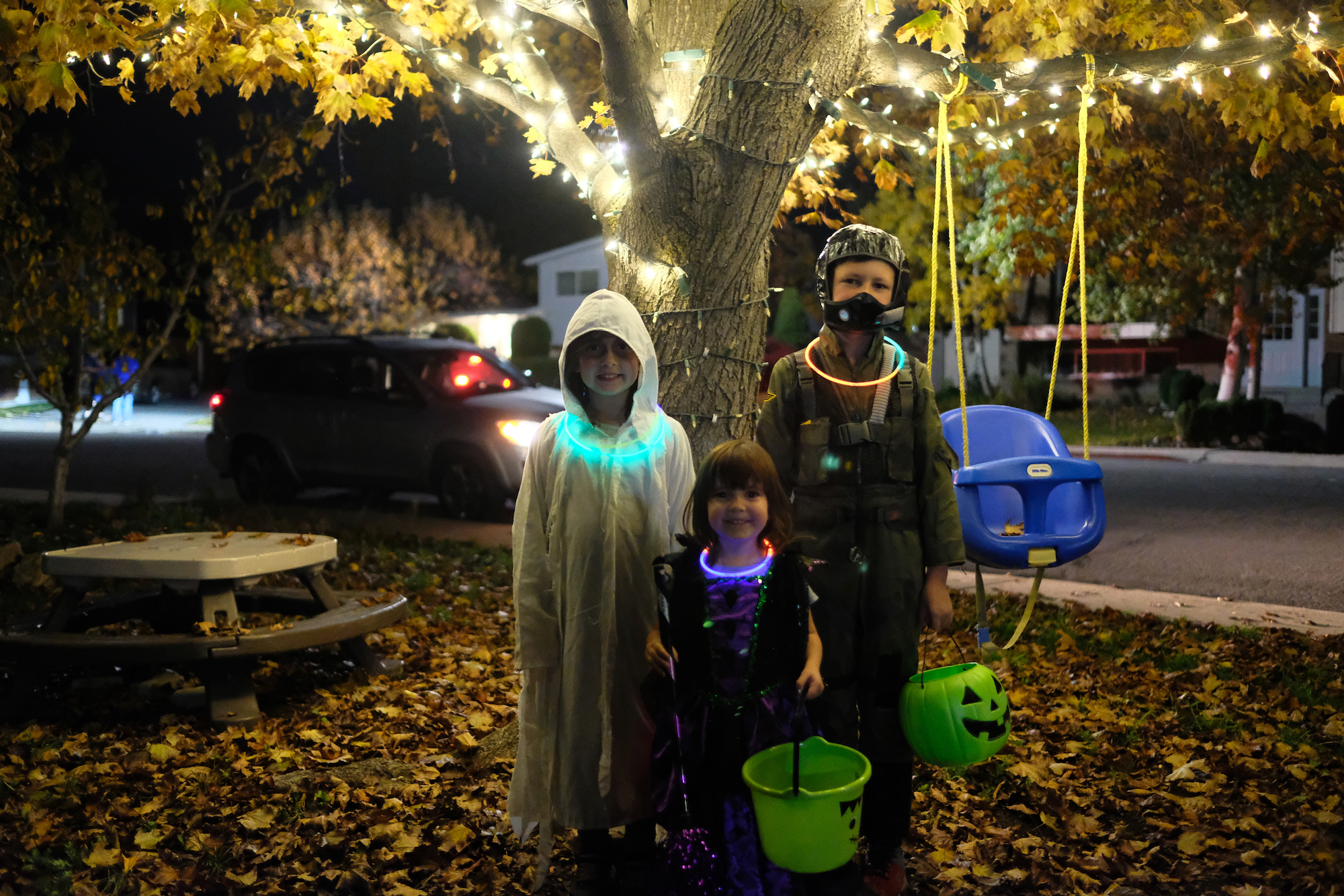Elle in a Ghost Costume, Emily as a witch, and Emmett in a Pilots costume standing under a lit tree on Halloween.